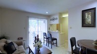 This single-floor residence offers a welcoming and functional layout designed for comfortable everyday living. The main living and dining area is bathed in natural light, courtesy of a large sliding glass door fitted with vertical blinds that opens to an outdoor area, providing a seamless connection between the interior and exterior. The space features light-toned carpeting and white walls that enhance the bright, airy ambiance throughout.

A peninsula-style breakfast bar separates the living and dining area from the kitchen, offering casual seating for two or more with dark bar stools that provide a subtle contrast to the neutral surroundings. The kitchen area is accessed through this open pass-through design, contributing to the overall sense of openness and flow.

Recessed lighting in the ceiling supplements the abundant natural light, ensuring the space remains well-lit throughout the day and evening. The textured ceiling adds a subtle architectural detail to the room. Decorative wall accents and tasteful furnishings complete the welcoming atmosphere, making this a practical and inviting space suited for both relaxed daily living and casual entertaining.
