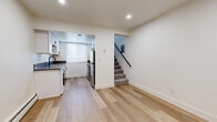This two-story residence offers a well-organized layout with thoughtfully updated interiors spread across both levels. The main floor features an open living area finished with light hardwood-style flooring that extends seamlessly toward the kitchen. The kitchen is designed in a galley configuration, outfitted with white shaker cabinetry accented by matte black hardware, gray countertops, a deep single-basin sink with a black faucet, and a full suite of stainless steel appliances including a gas range, microwave, and refrigerator. An in-unit washer is also conveniently integrated into the kitchen space. A window with white blinds allows natural light to brighten the area. Carpeted stairs with a sleek black handrail lead to the upper level, where bedrooms are finished with plush carpet flooring and include closets with built-in shelving. Baseboard heaters are present in multiple rooms. One room on the upper level provides direct access to an outdoor balcony, offering views of surrounding greenery. The bathroom is finished in a clean, all-white palette featuring large-format wall tiles, a soaking tub with a shower, matte black fixtures, a white vanity, and a small window for ventilation. Recessed lighting throughout the main level adds to the bright, modern ambiance of the space.