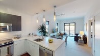 This contemporary single-floor residence showcases a thoughtfully designed open-concept layout that seamlessly connects the kitchen, dining, and living areas. The kitchen is a standout feature, equipped with stainless steel appliances including a refrigerator, range, and microwave, complemented by sleek white quartz countertops, a peninsula with an undermount sink, and a combination of white lower cabinetry and dark upper cabinets. Rich hardwood flooring grounds the kitchen space, while pendant lighting adds a refined touch overhead. The open dining area transitions naturally into the carpeted living room, which is bathed in natural light from multiple windows and a sliding glass door that opens to a private balcony with outdoor views. A track lighting system and additional pendant fixtures illuminate the shared living space. The bedroom is generously sized, featuring a large mirror, ample natural light, and access to a well-designed bathroom. The bathroom includes a floating vanity with dark wood cabinetry, a white countertop, a large mirror, and a tub-shower combination with tile flooring. Throughout the residence, crisp white walls and neutral tones create a bright, airy ambiance that is both modern and inviting.
