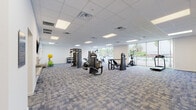 This single-floor fitness facility is designed to support a comprehensive range of exercise and wellness activities. The main workout area is generously proportioned, featuring a patterned blue-gray carpet tile floor, a suspended drop ceiling with recessed LED panel lighting, and crisp white walls that create a clean, professional atmosphere. Floor-to-ceiling windows along one side of the main gym floor flood the space with natural light while offering views of the surrounding greenery, enhancing the overall workout environment.

The main floor is equipped with a diverse selection of strength and cardio machines, including a multi-station cable system, selectorized strength machines, a chest press, an elliptical trainer, treadmills, stationary spin bikes, and free-weight benches paired with a dumbbell rack. The equipment is well-spaced, allowing for comfortable movement throughout the area.

A dedicated functional training room provides a bright, open, carpeted space stocked with stability balls, medicine balls, resistance bands, and foam rollers — ideal for group exercise, stretching, or personal training sessions. An additional flexible room, currently unfurnished, offers a versatile blank canvas suitable for yoga, classes, or other specialized programming.

The facility maintains a consistent modern aesthetic throughout, with uniform flooring, neutral walls, and efficient overhead lighting in every area.