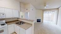 This single-floor residence offers an open, flowing layout that seamlessly connects the kitchen and living area, creating a cohesive and functional living space. The kitchen features white cabinetry, a double-basin sink, laminate countertops, and a breakfast bar peninsula that transitions naturally into the main living area — ideal for both everyday use and entertaining.

The living room is highlighted by soaring vaulted ceilings with elegant crown molding, adding a sense of height and grandeur to the space. A ceiling fan is centrally positioned, enhancing comfort and air circulation throughout. A classic fireplace with a white mantel serves as the focal point of the living area, providing both warmth and a charming architectural detail. Vertical blinds cover a large sliding glass door or window that invites an abundance of natural light into the room, brightening the neutral, warm-toned walls and plush carpeted floors.

The overall ambiance is warm and inviting, characterized by light neutral tones, clean lines, and thoughtful design details. The open sightlines between the kitchen and living areas enhance the sense of space, while the vaulted ceilings and natural light contribute to an airy, comfortable atmosphere throughout this well-appointed home.