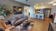 This single-floor residence showcases an open-concept layout that seamlessly connects the living area to the kitchen, creating a cohesive and functional living environment. The living space features warm hardwood-style flooring that extends throughout, complemented by neutral taupe-toned walls that lend a contemporary yet inviting atmosphere. Recessed lighting illuminates the space evenly, while a ceiling fan adds both style and practicality.

The living area comfortably accommodates a full-sized sofa and a round wooden coffee table set atop a textured area rug, with wall space utilized for decorative artwork, including a large world map print that serves as a focal point. An open archway leads directly to a dedicated laundry area, housing an in-unit washer and dryer — a convenient and practical feature.

The kitchen is positioned at the far end of the open layout and includes dark espresso-toned cabinetry, a granite-style countertop, stainless steel appliances, and a built-in microwave. A raised breakfast bar with bar stools provides additional seating and defines the transition between the kitchen and living areas, enhancing the space's overall flow and functionality.