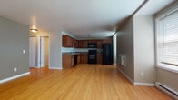 This single-floor unit offers a practical and well-organized layout, beginning with an open-concept main living area that seamlessly connects to the kitchen. The kitchen is fully equipped with warm-toned wooden cabinetry extending to the ceiling, granite-style countertops, and a full suite of black appliances, including a refrigerator, range, built-in microwave, and dishwasher. Light hardwood-style flooring runs throughout the main living and kitchen areas, contributing to a bright and cohesive feel. A window fitted with horizontal blinds allows natural light into the living space, which also features a wall-mounted air conditioning unit and baseboard heating for year-round climate control. A large sliding-door closet provides additional storage within the main area. The bedrooms are carpeted in a neutral gray tone and feature dedicated closets with built-in shelving and hanging rods. One bedroom benefits from a wide window with a baseboard heater beneath it. The bathroom includes large-format tile flooring, a tub-shower combination with built-in shelving, a toilet, and a wooden vanity with a white countertop. The overall color palette is neutral throughout, creating a clean and move-in-ready environment.