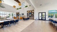 Located at 1900 E Inland Empire Blvd in Ontario, California, this single-floor community space offers a generously sized, open-concept layout spanning a substantial footprint. The design is defined by dramatically soaring ceilings with large black-framed clerestory windows that flood the interior with natural light, creating an airy and uplifting atmosphere throughout.

The entrance welcomes visitors with light wood-look flooring, oversized planters housing lush tropical trees, and floor-to-ceiling black-framed glass windows overlooking the exterior. The main gathering area transitions between elegant herringbone-patterned tile and wide-plank light wood flooring, visually delineating distinct functional zones. Seating arrangements include casual lounge groupings with cognac leather chairs flanking a fireplace, communal dining tables with navy and wood chairs, and soft seating areas anchored by landscape artwork and potted olive trees.

Built-in warm wood shelving units with integrated lighting serve as focal points, displaying curated décor alongside a wall-mounted television. Statement amber globe pendant lights hang from the high ceilings, adding warmth and sophistication. Black-framed glass double doors provide transitions between zones and lead to outdoor areas. The overall aesthetic is modern and refined, balancing bright whites with warm wood tones, greenery, and carefully selected furnishings.