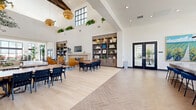 Located at 1900 E Inland Empire Blvd in Ontario, California, this single-floor community space offers a generously sized, open-concept layout spanning a substantial footprint. The design is defined by dramatically soaring ceilings with large black-framed clerestory windows that flood the interior with natural light, creating an airy and uplifting atmosphere throughout.

The entrance welcomes visitors with light wood-look flooring, oversized planters housing lush tropical trees, and floor-to-ceiling black-framed glass windows overlooking the exterior. The main gathering area transitions between elegant herringbone-patterned tile and wide-plank light wood flooring, visually delineating distinct functional zones. Seating arrangements include casual lounge groupings with cognac leather chairs flanking a fireplace, communal dining tables with navy and wood chairs, and soft seating areas anchored by landscape artwork and potted olive trees.

Built-in warm wood shelving units with integrated lighting serve as focal points, displaying curated décor alongside a wall-mounted television. Statement amber globe pendant lights hang from the high ceilings, adding warmth and sophistication. Black-framed glass double doors provide transitions between zones and lead to outdoor areas. The overall aesthetic is modern and refined, balancing bright whites with warm wood tones, greenery, and carefully selected furnishings.
