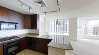 This single-floor residence offers a thoughtfully designed open-concept layout that seamlessly connects the kitchen and living areas. The kitchen is a standout feature, showcasing rich dark wood cabinetry that provides ample storage, complemented by elegant granite countertops with warm tonal variation. A peninsula counter extends the workspace and serves as a natural dividing element between the kitchen and the adjoining living space. The kitchen is equipped with a built-in dishwasher and a double-basin sink, and is illuminated by a combination of track lighting overhead and stylish pendant lights that hang above the peninsula, adding a modern touch. Flooring transitions from tile in the kitchen to plush carpet in the living area, offering both practicality and comfort. Large windows along the far wall flood the living space with generous natural light, creating a bright and airy ambiance throughout. The neutral, cream-toned walls enhance the sense of openness, while the overall design balances modern finishes with functional living. The space presents a clean, contemporary aesthetic well-suited for everyday living.