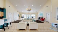 This single-floor community clubhouse offers a well-organized and inviting environment designed to serve a variety of resident needs. The main open area is defined by soaring vaulted ceilings, recessed lighting, and an elegant chandelier that anchors the central lounge space. Large windows and French doors along the perimeter flood the interior with abundant natural light, while ceiling fans contribute to a comfortable atmosphere throughout.

The lounge area is furnished with white upholstered seating arranged around a central coffee table, complemented by dark shag area rugs that add texture and warmth to the light-toned tile flooring. A business center section is integrated seamlessly into the open floor plan, featuring multiple computer workstations, black office chairs, and bold red accent seating, creating a functional workspace without sacrificing the room's airy feel.

A teal-accented snack bar counter with white bar stools offers a casual seating spot, and a dedicated kitchen area behind it features light wood cabinetry, a stainless steel refrigerator, a microwave, a coffee maker, and generous counter space — ideal for resident gatherings or events. Coastal-themed artwork adorns the warm cream-colored walls, reinforcing a relaxed, cheerful aesthetic. Additional doors lead to restrooms and staff-only areas, ensuring practical functionality throughout the space.