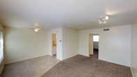 This single-floor residence offers a straightforward and functional layout designed for comfortable everyday living. The main living area is characterized by a combination of tile flooring near the entry and plush carpeting throughout the carpeted zones, creating a natural distinction between spaces. A ceiling fan with a light fixture is centrally positioned overhead, providing both airflow and ambient lighting, while a multi-bulb ceiling fixture in the adjacent area adds additional illumination.

The open layout connects the living space to surrounding rooms with ease. A doorway leads to a kitchen area, where cabinetry and countertops are visible, suggesting a practical and well-organized cooking space. A second doorway opens to what appears to be an additional room bathed in natural light from a nearby window, hinting at bright and airy secondary spaces.

The walls throughout are finished in a clean, neutral tone, complemented by a textured ceiling that adds subtle visual depth. HVAC vents are integrated into the walls, ensuring climate control throughout the space. The overall ambiance is calm and understated, offering a neutral canvas well-suited for a variety of interior styles and furnishing preferences.