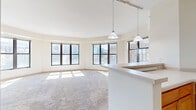 This single-floor urban unit is designed with an open, airy layout that makes excellent use of natural light. The living area is generously sized, featuring soft carpeting and a striking arrangement of large, black-framed windows along an angled wall that frame sweeping city skyline views. Pendant lights and a track lighting system hang overhead, adding a modern touch to the bright, neutral interior.

The kitchen is positioned adjacent to the living area and is separated by a peninsula counter, creating a functional open-concept flow. It is equipped with warm honey-toned wood cabinetry, white countertops, a white electric range, a dishwasher, and a double sink. Earthy stone-look tile flooring grounds the space, and additional windows provide natural light and city views.

The bedrooms are carpeted and well-lit, each benefiting from large windows that overlook the surrounding urban landscape. The rooms offer generous proportions with closet access and neutral wall tones throughout.

The bathroom includes a full tub-shower combination, a wood-toned vanity, and the same stone-look tile flooring found in the kitchen, maintaining a cohesive aesthetic. Overall, the unit presents a clean, functional design with consistent finishes and abundant natural light throughout.