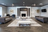 Valora is a thoughtfully designed single-floor commercial space that blends contemporary elegance with functional comfort. The centerpiece of the main lounge is a dramatic floor-to-ceiling fireplace surround crafted in textured stone and flanked by classical columns, with the "VALORA" name displayed prominently in gold lettering above — immediately establishing a strong brand identity upon entry.

Rich, dark hardwood flooring extends throughout the entire space, anchoring the light gray walls and dark ceiling that together create a sophisticated, upscale atmosphere. Recessed lighting and ceiling fans with integrated lighting provide a well-distributed, warm illumination across the open layout.

The main lounge area is generously proportioned, furnished with a plush channeled velvet sofa, accent chairs, and a marble-and-gold coffee table set atop a stylish area rug with gold veining details. A mounted flat-screen television and bar-height counter with seating provide additional functionality.

A secondary area features dual wall mirrors, sleek black leather accent chairs, and a workstation counter — ideal for consultations or client services. Arched architectural details and abundant natural light from large white-trimmed windows with horizontal blinds further enhance the refined, welcoming ambiance throughout the space.