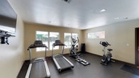 This single-floor fitness center is thoughtfully designed to support a comprehensive workout experience. The space features a clean, functional layout with neutral beige walls and a textured white ceiling fitted with recessed lighting that ensures bright, even illumination throughout. Durable dark rubber flooring provides both safety and comfort underfoot, making it well-suited for high-impact exercise. The cardio area is equipped with multiple treadmills and an elliptical trainer, offering a range of options for aerobic workouts. A stationary bike is also available for low-impact cycling sessions. A dedicated free weight rack with a selection of dumbbells rounds out the strength training options. Large windows along the front wall allow generous natural light to fill the space and offer views of the outdoor surroundings, including a glimpse of an adjacent outdoor area with a fence and landscaping. A smaller window on the side wall adds additional ventilation and light. The overall ambiance is clean, practical, and inviting, creating a motivating environment for residents or users to pursue their fitness goals.