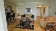 This single-story space presents a warm and inviting living area finished with light hardwood flooring that extends throughout the main areas. The living room serves as the central gathering space, featuring classic crown molding that adds a touch of architectural refinement to the neutral-toned walls. A beige sofa accented with dark decorative pillows anchors the seating area, complemented by a dark wood coffee table with drawer storage resting atop a bold floral-patterned area rug. A framed wall mirror adds depth and visual interest to the space. Natural light filters in, enhancing the open and airy feel of the room. A bar-height seating area with dark chairs is visible along one side, offering a casual dining or counter space option. A hallway extends toward what appears to be additional rooms, including a bedroom visible through a doorway, where light-colored furnishings continue the cohesive, understated aesthetic. The entry area features a flush-mount light fixture and a solid front door, providing a defined and functional entryway. Overall, the space combines practical layout with classic design touches.