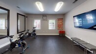This single-floor fitness room is thoughtfully designed to support a well-rounded exercise routine. The space features dark, cushioned rubber flooring throughout, providing both safety and comfort during workouts. Bright overhead fluorescent lighting ensures the room is well-illuminated at all times, while multiple windows along one wall allow natural light to filter in and offer views of the surrounding exterior.

The room is organized into functional zones. A dedicated cycling area houses several stationary spin bikes positioned in front of a pair of large, dark-framed wall mirrors — ideal for monitoring form during rides. Motivational wall decals, including an "Inhale. Exhale." design, contribute to the focused ambiance of the space.

On the opposite end of the room, a collection of red stability exercise balls, foam mats, and storage containers provide resources for stretching and floor exercises. A modular gray shelving unit offers organized storage for smaller equipment and accessories. A wall-mounted flat-screen television displays fitness app content, adding a modern, tech-integrated element to the space.

A padded bench provides a convenient spot for rest or stretching. The clean, neutral gray walls and consistent design aesthetic throughout give the room a professional yet welcoming atmosphere, making it a functional and motivating environment for fitness activities.