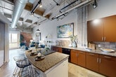 Located at 1 Hermann Park Court in Houston, TX, this two-bedroom, two-bathroom loft showcases a striking industrial-chic aesthetic with soaring ceilings, exposed concrete beams, visible ductwork, and raw brick accent walls that lend the space a bold, urban character. The open-concept layout seamlessly connects the kitchen, dining, and living areas into one dynamic, light-filled space anchored by polished concrete floors throughout.

The kitchen is well-appointed with rich wood cabinetry, granite countertops, a tile backsplash, black appliances, and a central island with bar seating — ideal for both cooking and entertaining. The adjacent living and dining area benefits from the soaring ceiling height and dramatic exposed structural elements, creating an expansive, loft-style atmosphere.

The primary bedroom features an exposed brick accent wall, a large window with sweeping views of Hermann Park and the Houston skyline, and a concrete ceiling that echoes the loft's industrial roots. The second bedroom offers a quieter, refined aesthetic with elevated ceilings and pendant lighting. Both bathrooms include granite vanities and tile details, with one featuring a walk-in shower and the other a shower-tub combination. A dedicated laundry area and multiple closets add practical functionality to this architecturally distinctive home.