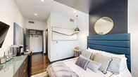 Located at 5151 S Rio Grande St in Littleton, CO, this thoughtfully designed one-bedroom, one-bathroom unit offers a compact yet well-appointed layout across a single floor. The bedroom serves as the centerpiece of the space, showcasing warm hardwood flooring, crisp white walls, and a striking navy blue accent wall that adds bold contrast and depth. A tufted upholstered headboard in matching navy anchors the room, complemented by dual pendant lights with matte black shades that provide both function and style. A gold-framed circular mirror mounted on the accent wall adds an elegant finishing touch. Recessed lighting and a wall-mounted flat-screen TV round out the room's modern amenities, while a window allows natural light to filter in. A walk-in closet provides practical storage just off the main sleeping area. The bathroom continues the contemporary aesthetic with hexagonal tile flooring, a white vanity with clean cabinet hardware, and decorative wall art that ties the design together. The overall ambiance of the space is modern and curated, blending neutral tones with bold accents to create a polished, comfortable living environment ideal for those seeking stylish, efficient living in the Littleton area.