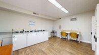 Located at 1526 Brookdale Ave in Cookeville, Tennessee, this dedicated laundry facility serves the Hampton Creek community. The single-room space is thoughtfully designed to accommodate residents' laundry needs efficiently and comfortably.

The room is lined along one wall with a row of front-load washing machines, complemented by stacked dryer units nearby, providing ample laundry capacity. A utility sink with a stainless steel trash receptacle is conveniently positioned adjacent to the machines. A built-in folding counter with two wicker chairs offers a practical surface for sorting and folding laundry, while an additional small table with two wooden chairs near the entrance provides a comfortable waiting area for residents.

The interior features warm wood-look flooring throughout, white walls, and clean white trim, contributing to a bright and tidy atmosphere. Overhead fluorescent lighting ensures the space is well-lit, and a large skylight panel further enhances illumination. Natural light also enters through double windows fitted with white blinds. The facility includes a wall-mounted paper towel dispenser, posted laundry center rules, and a fire extinguisher for safety. A glass entry door with the Hampton Creek branding completes the welcoming, well-maintained environment.