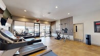 This fitness center is a thoughtfully designed, single-floor facility offering a comprehensive range of exercise options in a modern, welcoming environment. The main workout area is anchored by polished concrete flooring in a warm amber tone, which lends the space a clean yet inviting aesthetic. Recessed lighting throughout ensures consistent, well-lit conditions for all activities.

The cardio zone is lined with True-brand treadmills, an elliptical trainer, and a rowing machine, all positioned to take advantage of large floor-to-ceiling windows and glass doors that flood the room with natural light and provide views of an outdoor pool and terrace. A wall-mounted flat-screen TV and wall mirrors add to the functionality of this area, while a bold green accent wall contributes a dynamic visual element.

Strength and functional training equipment, including a multi-station cable machine, a captain's chair, and a stationary bike, are arranged along a feature wall adorned with vertical wood slat paneling and a motivational "Workout" graphic. Fitness reference charts are posted throughout for guidance.

A separate yoga and stretching room offers a serene retreat, featuring ornate mandala floor rugs, full-length mirrors with ballet-style bars, a wall-mounted TV, live plant accents, and a dedicated mini-split HVAC unit for personalized climate control. The space is designed to support both high-intensity training and mindful movement.