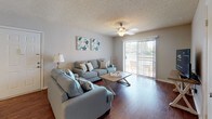 This single-floor residence offers a thoughtfully designed layout that balances comfort and functionality throughout. The living room serves as the heart of the home, featuring rich hardwood flooring, a ceiling fan, and a large sliding glass door that opens to an outdoor balcony, filling the space with natural light and providing a seamless indoor-outdoor connection. Adjacent to the living area, the dining space accommodates a round table beneath an elegant chandelier, complemented by a wall-mounted mirror that enhances the sense of openness. The galley-style kitchen is efficiently arranged with gray cabinetry, granite-style countertops, a tile backsplash, and a full suite of appliances including a stainless steel dishwasher, black refrigerator, and electric range. A peninsula counter connects the kitchen to the main living area. The primary bedroom features plush carpeting, a window with natural light, and ample space for a dresser and full-size bed. The secondary bedroom is equally comfortable with carpeted floors and a bright window. The bathroom includes a granite-style vanity countertop and a shower-tub combination. Textured ceilings are consistent throughout, adding a classic character to the overall aesthetic.