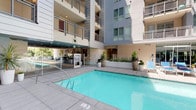 Located at 201 W Lexington Dr in Glendale, California, this outdoor amenity space centers around a generously proportioned swimming pool set within a striking modern courtyard. The pool area is enclosed by a multi-story residential building whose contemporary architecture — characterized by clean lines, neutral facades, metal-railed balconies, and large glass windows — frames the space on all sides, creating a private, resort-like atmosphere open to the Southern California sky.

The pool itself features clear turquoise water and a wide surrounding deck finished in smooth concrete pavers, providing ample room for lounging. Blue chaise lounges are arranged along one side of the pool beneath shade umbrellas, offering a comfortable retreat for relaxation. Lush potted plants and tropical greenery are strategically placed throughout the courtyard, softening the modern architectural elements and adding a vibrant, welcoming touch.

Accessible restroom facilities are conveniently located poolside, along with an elevator entrance that connects the courtyard to the broader building. Glass-paneled doors provide interior access, and the well-maintained surroundings reflect a high standard of upkeep. The space benefits from abundant natural light throughout the day, making it an inviting outdoor destination year-round in Glendale's mild climate.