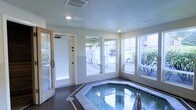 This single-floor indoor spa room is designed as a dedicated relaxation and wellness space. At its center sits an octagonal hot tub finished with decorative tile surround and coping, set into a dark tile floor that adds contrast to the room's light, neutral-toned walls. The space is well-lit by recessed ceiling lights, complemented by abundant natural light streaming through multiple large windows along two walls. These windows offer views of the surrounding outdoor area, including greenery and a parking or courtyard area, creating a bright and open atmosphere despite the enclosed setting. A textured ceiling with HVAC vents ensures climate control within the room. To the left, a glass-panel door — likely leading to a sauna or changing area — adds additional functionality to the wellness suite. A separate exterior door with a wooden frame provides direct outdoor access, enhancing convenience and ventilation. The overall design prioritizes practicality and comfort, with clean finishes and a straightforward layout suited to a shared or private amenity space within a larger residential or hospitality property.