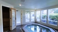 This single-floor indoor spa room is designed as a dedicated relaxation and wellness space. At its center sits an octagonal hot tub finished with decorative tile surround and coping, set into a dark tile floor that adds contrast to the room's light, neutral-toned walls. The space is well-lit by recessed ceiling lights, complemented by abundant natural light streaming through multiple large windows along two walls. These windows offer views of the surrounding outdoor area, including greenery and a parking or courtyard area, creating a bright and open atmosphere despite the enclosed setting. A textured ceiling with HVAC vents ensures climate control within the room. To the left, a glass-panel door — likely leading to a sauna or changing area — adds additional functionality to the wellness suite. A separate exterior door with a wooden frame provides direct outdoor access, enhancing convenience and ventilation. The overall design prioritizes practicality and comfort, with clean finishes and a straightforward layout suited to a shared or private amenity space within a larger residential or hospitality property.
