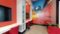 This single-floor commercial space makes an immediate impression with its bold, automotive-inspired design. The interior is defined by vivid red walls that energize the environment and set a dynamic, brand-forward tone throughout. A standout feature is the dramatic full-wall photographic mural depicting a high-speed car in motion, complete with vivid streaks of light and a close-up of a vehicle's side mirror — an eye-catching focal point that anchors the room's theme.

A built-in white shelving unit runs vertically along one wall, offering organized display space for products or branded accessories. Adjacent to it, a wall-mounted flat-screen television is integrated into a recessed niche, providing a functional media element for customer engagement or informational display.

A continuous white counter runs along the wall beneath the shelving and screen, accompanied by sleek red pedestal stools that complement the overall color scheme. Recessed ceiling lighting provides focused illumination throughout the space, enhancing both functionality and ambiance. The floor is finished with a neutral, textured carpet tile that grounds the bold palette above. The overall design is cohesive, modern, and clearly tailored to reflect an automotive brand identity, making it well-suited for use as a customer lounge, showroom waiting area, or service center lobby.