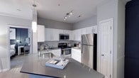 This single-floor space showcases a well-appointed kitchen designed with a clean, contemporary aesthetic. White shaker-style cabinetry lines the walls, providing ample storage while contributing to the bright, open feel of the room. The cabinetry is complemented by a classic subway tile backsplash that adds texture and visual interest without overwhelming the space.

The kitchen is equipped with a full suite of stainless steel appliances, including a gas range, built-in microwave, and a full-sized refrigerator. A track lighting fixture overhead ensures consistent illumination throughout the cooking area, while a stylish cylindrical pendant light hangs above the kitchen island, adding a decorative focal point.

The expansive island features a smooth gray countertop that extends generously, offering both additional workspace and a casual dining or entertaining surface. An undermount sink with a modern arc-neck faucet is integrated into the island, adding functionality. The gray and white color palette throughout creates a cohesive, polished atmosphere that feels both modern and inviting.