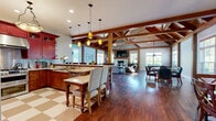 Located at 380 Mather St in Hamden, Connecticut, this single-floor space makes a striking impression through a combination of rustic craftsmanship and thoughtful design. The living room is the architectural centerpiece, showcasing dramatic cathedral ceilings with exposed timber beam trusses and a commanding floor-to-ceiling stone fireplace. Rich hardwood flooring runs throughout, anchoring the warm, lodge-inspired aesthetic. Abundant natural light filters through multiple windows fitted with wooden shutters, enhancing the inviting ambiance. The kitchen is a bold and functional space defined by deep red cabinetry, stainless steel appliances, granite countertops, and a checkered tile floor. A generously sized island with bar seating serves as a natural gathering point, illuminated by a curated arrangement of pendant lights. The dining area offers an elegant yet relaxed setting, centered around a substantial wood farmhouse table beneath a geometric lantern-style chandelier. Wall mirrors and vibrant artwork add dimension and personality to the space. French doors in the dining area provide a seamless visual connection to adjacent areas. Together, these rooms create a cohesive, character-filled environment that balances rustic warmth with refined detail.