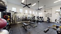 This fitness center is designed to support a full range of workout routines within a spacious, single-story layout. The room features notably high ceilings adorned with crown molding and multiple large ceiling fans, contributing to an open, airy atmosphere. Rows of multi-pane windows line the walls at both standard and clerestory heights, flooding the interior with natural light and offering views of the surrounding outdoor landscape.

The flooring throughout is covered in durable dark rubber tiles, providing a safe and impact-absorbing surface ideal for strength and cardio training. Large wall-mounted mirrors extend along one side of the room, visually expanding the space while allowing users to monitor their form during exercise.

The cardio zone is well-appointed with multiple treadmills, elliptical trainers, and stationary bikes. The strength training area includes adjustable weight benches, a full dumbbell rack, cable machines, and various selectorized resistance machines to support comprehensive muscle group training. A dedicated functional fitness corner houses medicine balls, stability balls, and additional accessories.

Mounted flat-screen televisions are positioned for visibility throughout the room. The covered outdoor patio area, framed by natural stone columns and exposed beam accents, provides an inviting transition between the interior fitness space and the exterior surroundings.
