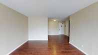 This single-floor space offers a straightforward and functional layout designed for comfortable living. The main living area is open and airy, featuring rich, dark hardwood-style flooring that runs continuously throughout, adding warmth and visual depth to the space. The walls are finished in a soft, neutral beige tone, creating a clean and versatile backdrop that pairs well with a variety of furnishings and décor styles. White baseboards provide a crisp contrast against both the flooring and the walls, contributing to a polished overall aesthetic. The ceiling is smooth and white, fitted with a flush-mount light fixture that provides practical illumination to the room. A sliding door on one side of the living area offers access to additional storage or a closet, adding to the unit's functionality. The entryway is clearly defined, featuring a solid white door with standard hardware, and is enhanced by a wall-mounted security panel, offering a sense of safety and convenience. The open flow between the entryway and living area creates a seamless transition throughout the space. Overall, the unit presents a clean, move-in-ready environment with practical finishes suited for a range of living arrangements.