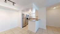 This single-floor residence features a clean, light-filled interior defined by crisp white walls, textured ceilings, and large-format cream tile flooring that extends throughout the main living areas. The kitchen is efficiently designed with white cabinetry, dark granite-style countertops, and a full suite of stainless steel appliances including a refrigerator, microwave, and range. A curved peninsula provides additional counter space and separates the kitchen from the open living area. Track lighting and a pendant cluster illuminate the kitchen, while a ceiling fan serves the spacious main living area, which benefits from natural light through a large window and direct exterior access via a glass-paneled door. A secondary living or dining zone flows seamlessly from the kitchen, offering additional flexible space. The bedroom features plush carpeting, a ceiling fan, a built-in closet with louvered doors, and direct access to a private en-suite bathroom. The bathroom is fitted with a white vanity topped with a granite-style surface, a mirrored medicine cabinet, and a full tiled tub-shower combination, all finished in a clean, neutral palette throughout.