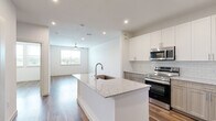 This thoughtfully designed single-floor unit showcases a clean, contemporary aesthetic defined by crisp white walls, rich hardwood-style flooring, and recessed lighting throughout. The open-concept layout seamlessly connects the kitchen and living area, creating a spacious and cohesive flow. The kitchen is a standout feature, anchored by a large center island topped with granite countertops and an undermount sink. White upper cabinetry contrasts elegantly with warm wood-toned lower cabinets, while a subway tile backsplash adds a refined touch. Stainless steel appliances — including a French door refrigerator with ice maker, electric range, over-the-range microwave, and dishwasher — complete the well-appointed cooking space. The living area is bright and airy, benefiting from multiple windows that frame outdoor views and a ceiling fan that enhances comfort. Additional rooms offer versatile spaces bathed in natural light through window-blind-fitted windows. The bathroom is finished with matching granite countertops, warm wood-toned cabinetry, a tiled soaking tub and shower, and direct access to a walk-in closet with built-in shelving. A dedicated laundry area is also visible within the unit, adding everyday convenience to this polished, modern space.
