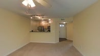 This single-floor residence is designed with an open-concept layout that connects the living area and kitchen, creating a cohesive and functional living space. The living area is finished with wall-to-wall carpeting in a neutral tone, contributing to a warm and comfortable atmosphere. A ceiling fan with an integrated light fixture is prominently positioned overhead, providing both air circulation and ambient lighting throughout the space.

The kitchen is positioned at the rear of the living area and is accessible via a pass-through counter with a dark countertop, which also serves as a casual dining or prep surface. Light wood cabinetry lines the kitchen walls, complemented by stainless steel appliances including a refrigerator and range. Track lighting mounted on the kitchen ceiling provides focused task illumination.

A hallway visible from the living area leads to the front entry door, which features small decorative windows that allow natural light to filter into the space. The overall color palette consists of soft, warm yellows and neutral tones throughout the walls and flooring, lending the space a welcoming and understated character. The layout is practical and efficiently organized for everyday living.