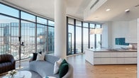 Perched high above the city, this contemporary residence offers a striking living experience defined by expansive floor-to-ceiling glass walls that frame panoramic skyline views from virtually every room. Light wood flooring runs throughout, complementing the clean, white interior palette and lending warmth to the modern aesthetic.

The open-plan living and dining area is anchored by a graceful curved sofa and a sculptural pendant light, with dramatic wraparound glazing inviting abundant natural light. Cylindrical structural columns add an architectural punctuation to the space. The adjacent gourmet kitchen features high-gloss white cabinetry, a stone-slab backsplash, granite countertops, and premium built-in appliances including a gas cooktop and double wall ovens.

The bedrooms offer serene retreats, each benefiting from floor-to-ceiling windows that showcase impressive city vistas. Circular porthole-style windows in the hallway area add a distinctive design detail. The bathrooms are finished in marble tile, complete with floating white vanities, stone countertops, and illuminated mirrors, delivering a refined, hotel-like atmosphere. Throughout, the residence balances sophisticated minimalism with warm, curated details for an elevated urban living experience.