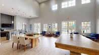 Located at Falcon Parc Blvd in Orlando, Florida, this single-floor community clubroom offers a welcoming and well-appointed gathering space designed for both casual socializing and organized events. The most striking architectural feature is the dramatically soaring ceiling height, which lends an airy, expansive feel to the interior well beyond what the footprint alone might suggest. Rows of tall, grid-paned French doors and clerestory windows line the walls, flooding the space with abundant natural light and offering views of the outdoor amenity area beyond.

The main open area is furnished with a large rustic wooden communal table as its centerpiece, complemented by additional seating arrangements including a modern dining table with upholstered chairs and intimate lounge groupings featuring blue accent chairs. Light wood-look flooring runs throughout, enhancing the bright and clean aesthetic defined by soft gray walls.

A dedicated kitchenette is tucked to one side, equipped with dark wood cabinetry, granite countertops, a stainless steel refrigerator, a double sink, and a breakfast bar with bar seating — making it well-suited for catered events or community gatherings. A separate flex room accessible through floor-to-ceiling glass French doors features a wall-mounted television, offering a more private setting within the overall space.