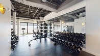 Located at 161 W 600 S in Salt Lake City, Utah, this expansive fitness center is designed to accommodate a full range of workout styles within a single, open-plan floor. The space features an industrial-inspired aesthetic, with exposed ceilings that reveal structural trusses, ductwork, and mechanical systems overhead, lending the facility a modern, urban character. Durable rubber flooring spans the workout areas, providing both safety and functionality throughout.

The strength training zone is well-appointed with multiple power racks loaded with weight plates, a comprehensive rack of dumbbells ranging across various weights, and adjustable benches — all positioned alongside full-length mirrors that enhance spatial depth and allow users to monitor their form. A dedicated functional training area includes TRX suspension systems, battle ropes, plyo boxes, kettlebells, medicine balls, and cable machines, offering versatile options for dynamic and cross-training workouts.

The cardio and selectorized machine section benefits from floor-to-ceiling windows that frame sweeping views of the Salt Lake City skyline and the surrounding Wasatch mountain range, flooding the space with natural light. Structural columns punctuate the open layout, and the generous ceiling height contributes to an airy, unrestrictive atmosphere throughout the facility.