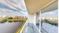 This unit includes a private balcony that serves as a notable outdoor feature, elevated above the surrounding landscape to provide open, unobstructed views of a tranquil pond and manicured green space below. The balcony is finished with a smooth concrete floor and enclosed by a white metal railing, offering both safety and an unimpeded sightline across the water. Outdoor seating, including a glass-top table and metal chairs, is positioned to take full advantage of the pleasant surroundings, making it a functional space for relaxation or casual outdoor dining.

Access to the interior is provided through a full-width sliding glass door system, which allows abundant natural light to flow between the balcony and the living area within. The textured stucco ceiling overhead is consistent with the building's exterior construction. Neighboring residential buildings and clusters of palm trees are visible in the distance, providing context for the community setting. The overall ambiance of the balcony is calm and airy, with open sky views and natural waterfront scenery creating a peaceful outdoor environment well-suited for enjoying the surrounding Florida-style landscape.