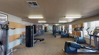 This single-floor fitness center offers a spacious and well-organized workout environment designed to support a full range of exercise routines. The room is distinguished by its bold, horizontally striped orange and white walls, giving the space an energetic and motivating ambiance. Durable, patterned carpet tiles cover the floor throughout, providing a practical and comfortable surface underfoot.

The cardio section is generously appointed with multiple treadmills, elliptical trainers, and a stationary bike, all positioned near large windows that allow natural light to fill the space and offer views of the surrounding outdoor area. The strength training section features a variety of cable and weight machines, including a lat pulldown, leg press, and multi-station cable system, as well as a dedicated bench press setup. A tall, freestanding unit — likely an infrared sauna or massage pod — adds an additional wellness element to the facility.

Overhead fluorescent lighting ensures the entire space is brightly and evenly lit. A large mirror along one wall enhances the sense of openness and allows users to monitor their form during workouts. Informational signage and framed artwork are mounted on the walls, contributing to a polished, well-maintained atmosphere. The layout is thoughtfully arranged to allow ample circulation between equipment.