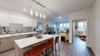 This well-appointed residence offers a thoughtfully designed single-story layout encompassing two bedrooms and two bathrooms. The open-concept living area seamlessly connects the kitchen, dining, and living spaces, creating a fluid and functional environment ideal for both everyday living and entertaining.

The kitchen is a standout feature, showcasing an expansive island with a white quartz countertop and seating for multiple guests, complemented by a series of modern pendant lights overhead. Cabinetry combines white upper cabinets with warm wood-toned lower cabinets, while a mirrored backsplash amplifies light throughout the space. Stainless steel appliances, including a range with a wall-mounted hood vent and a French door refrigerator, complete the chef-inspired kitchen.

The living room receives generous natural light through large windows that frame views of the surrounding neighborhood. Wide-plank hardwood-style flooring runs consistently throughout the unit, lending a cohesive, contemporary feel.

The spacious primary bedroom features large windows and ample room, accompanied by a dedicated walk-in closet with built-in racking. The second bedroom also benefits from natural light and exterior views. Both bathrooms offer clean, modern finishes with wood-toned vanities, white quartz countertops, and large mirrors. A utility room provides additional practical storage space.