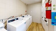 Located at 801 Chinquapin Rd in Columbia, South Carolina, this shared laundry facility is a single-room space designed for efficient, communal use. The room is constructed with painted concrete block walls and finished with durable wood-look vinyl plank flooring, giving it a clean and functional aesthetic.

The laundry area is well-equipped with a row of top-loading washing machines along one wall, each featuring digital control panels and coin-operated payment interfaces for convenient, self-service use. Alongside the washers, a bank of stacked dryer units lines the adjacent wall, maximizing the use of vertical space and accommodating multiple users simultaneously.

A built-in folding table with a marble-style surface provides a dedicated workspace for sorting and folding laundry. A window on one wall allows for natural light to enter the room while offering a view of the surrounding parking area. The space is also equipped with visible plumbing and utility infrastructure, an electrical panel, a fire extinguisher for safety compliance, and a security camera, reflecting a commitment to both safety and oversight. A keyed exterior door provides secure, controlled access to the facility. The overall layout is straightforward and practical, making it a convenient shared amenity.