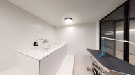 This single-level pet grooming room is thoughtfully designed to accommodate all aspects of pet care in a clean, modern environment. The space is defined by its crisp white large-format wall tiles, which line the washing area from floor to ceiling, creating a hygienic and easy-to-maintain surface. A raised, built-in pet wash basin dominates one side of the room, equipped with a wall-mounted faucet and handheld spray hose, alongside a tiled step for convenient pet access. The basin's white solid-surface construction integrates seamlessly with the surrounding tilework. Adjacent to the wash station sits a professional-grade grooming table with a grey non-slip surface and blue restraint loops, mounted on a wheeled, adjustable base for versatility. Sleek black-framed floor-to-ceiling glass partitions divide the grooming area from the adjoining corridor, allowing visual connectivity while maintaining separation. Light-toned tile flooring extends throughout, complementing the bright, neutral palette. Overhead, a flush-mount ceiling fixture provides even illumination across the grooming zone. The overall design balances practicality with a contemporary aesthetic, making it a well-equipped and visually cohesive pet care facility.