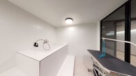This single-level pet grooming room is thoughtfully designed to accommodate all aspects of pet care in a clean, modern environment. The space is defined by its crisp white large-format wall tiles, which line the washing area from floor to ceiling, creating a hygienic and easy-to-maintain surface. A raised, built-in pet wash basin dominates one side of the room, equipped with a wall-mounted faucet and handheld spray hose, alongside a tiled step for convenient pet access. The basin's white solid-surface construction integrates seamlessly with the surrounding tilework. Adjacent to the wash station sits a professional-grade grooming table with a grey non-slip surface and blue restraint loops, mounted on a wheeled, adjustable base for versatility. Sleek black-framed floor-to-ceiling glass partitions divide the grooming area from the adjoining corridor, allowing visual connectivity while maintaining separation. Light-toned tile flooring extends throughout, complementing the bright, neutral palette. Overhead, a flush-mount ceiling fixture provides even illumination across the grooming zone. The overall design balances practicality with a contemporary aesthetic, making it a well-equipped and visually cohesive pet care facility.