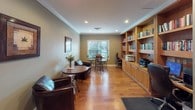 This single-floor space is designed as a multifunctional room combining lounge, workspace, and reading areas into one cohesive environment. The interior is unified by warm hardwood flooring, sage green walls, and crisp white crown molding that runs throughout, lending the space a polished yet comfortable character. Recessed lighting provides consistent illumination across the room.

The lounge area is furnished with dark leather chairs arranged around a round wooden bistro table accented with a floral centerpiece, creating an inviting setting for casual meetings or relaxation. Framed botanical artwork adorns the walls, contributing to the space's refined ambiance.

A built-in wooden bookcase lines one wall, filled with an extensive collection of books and housing a dedicated workstation equipped with a computer, keyboard, and office chair. Adjacent to the workstation, a printer and additional office equipment are neatly integrated into the cabinetry.

A separate dining nook features a round wooden table with matching chairs, positioned beside a large window fitted with horizontal blinds that filter natural light and offer views of surrounding greenery. The overall atmosphere is warm, organized, and well-appointed, suitable for both productivity and leisure.