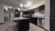 This single-story residence showcases a cohesive modern design highlighted by dark espresso cabinetry, granite countertops, and light wood-look flooring that flows throughout the main living areas. The kitchen serves as a central feature, equipped with a full suite of stainless steel appliances including a refrigerator, electric range, built-in microwave, and dishwasher. A large center island with a granite top provides additional workspace and casual seating, while pendant lighting overhead adds a refined touch. A distinctive diamond-pattern wood wine rack built into the upper cabinetry adds a unique architectural detail. The open layout connects the kitchen seamlessly to the living area, which features a ceiling fan, neutral walls, and a front entry door with access to natural light. Bedrooms are carpeted for comfort and include windows with blinds that allow ample natural light. Two bathrooms serve the unit: one features a glass-enclosed walk-in shower, while the other includes a full soaking tub and shower combination, both appointed with dark vanity cabinetry and granite countertops consistent with the overall design palette.