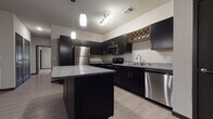 This single-story residence showcases a cohesive modern design highlighted by dark espresso cabinetry, granite countertops, and light wood-look flooring that flows throughout the main living areas. The kitchen serves as a central feature, equipped with a full suite of stainless steel appliances including a refrigerator, electric range, built-in microwave, and dishwasher. A large center island with a granite top provides additional workspace and casual seating, while pendant lighting overhead adds a refined touch. A distinctive diamond-pattern wood wine rack built into the upper cabinetry adds a unique architectural detail. The open layout connects the kitchen seamlessly to the living area, which features a ceiling fan, neutral walls, and a front entry door with access to natural light. Bedrooms are carpeted for comfort and include windows with blinds that allow ample natural light. Two bathrooms serve the unit: one features a glass-enclosed walk-in shower, while the other includes a full soaking tub and shower combination, both appointed with dark vanity cabinetry and granite countertops consistent with the overall design palette.