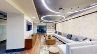 This expansive single-floor amenity space is designed with a distinctly modern aesthetic, offering multiple interconnected zones that balance social gathering with intimate retreat. The centerpiece of the main lounge is a striking ceiling installation featuring large circular LED light rings set against a dark recessed panel, which floats above warm hardwood flooring and a generously sized sectional sofa backed by a textured stone-clad accent wall. A walnut media console and open shelving unit with brass hardware add refined storage and display elements nearby.

Adjacent areas transition seamlessly through open corridors, where large-format slate tile flooring takes over and dining tables flanked by green velvet chairs create a distinct gathering space. A glass-enclosed private lounge room features its own curated seating, a wine display, and decorative artwork, accessible through sliding glass doors framed by herringbone-patterned tile at the threshold.

Further into the space, a game and lounge area offers plush seating in rich teal and cognac tones, built-in shelving with curated décor, and bold abstract artwork. A secondary corridor leads to an additional seating alcove brightened by natural light from a window. Throughout, the design integrates layered lighting, high-end furnishings, and cohesive color palettes to create a polished, hospitality-inspired environment.
