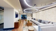 This expansive single-floor amenity space is designed with a distinctly modern aesthetic, offering multiple interconnected zones that balance social gathering with intimate retreat. The centerpiece of the main lounge is a striking ceiling installation featuring large circular LED light rings set against a dark recessed panel, which floats above warm hardwood flooring and a generously sized sectional sofa backed by a textured stone-clad accent wall. A walnut media console and open shelving unit with brass hardware add refined storage and display elements nearby.

Adjacent areas transition seamlessly through open corridors, where large-format slate tile flooring takes over and dining tables flanked by green velvet chairs create a distinct gathering space. A glass-enclosed private lounge room features its own curated seating, a wine display, and decorative artwork, accessible through sliding glass doors framed by herringbone-patterned tile at the threshold.

Further into the space, a game and lounge area offers plush seating in rich teal and cognac tones, built-in shelving with curated décor, and bold abstract artwork. A secondary corridor leads to an additional seating alcove brightened by natural light from a window. Throughout, the design integrates layered lighting, high-end furnishings, and cohesive color palettes to create a polished, hospitality-inspired environment.