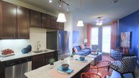 This single-floor space showcases a thoughtfully designed open-concept layout that connects the kitchen and living area into one cohesive, functional environment. The kitchen is outfitted with rich dark wood cabinetry, a light granite countertop island with bar seating, stainless steel appliances including a full-size refrigerator, a tile backsplash, and a undermount sink — all illuminated by modern pendant lighting and track lighting overhead. The kitchen island serves as a natural transition point into the dining and living areas, making it ideal for entertaining. The living room is warm and inviting, featuring a ceiling fan with integrated lighting, large windows dressed with coral-toned curtains that allow an abundance of natural light to fill the space, and views to the outdoors. The interior design is contemporary and vibrant, with bold accent colors including coral, blue, and teal complementing the neutral wall tones. Decorative touches such as potted succulents, patterned textiles, and layered area rugs contribute to an energetic yet comfortable ambiance throughout the space.