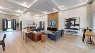 Located at 70 Lynn Rd in Durham, NC, this single-floor community amenity space offers a thoughtfully designed layout spanning a generous footprint. The heart of the space is a combined dining and living room characterized by soaring tray ceilings, recessed lighting, and wide-plank light hardwood flooring that runs throughout. The living area is anchored by a striking textured stone accent wall housing a mounted flat-screen TV, complemented by plush seating arrangements in rich leather and fabric tones. Large windows invite natural light and provide views of the surrounding greenery. The dining area accommodates multiple seating groups, with pendant lighting overhead and a kitchenette bar featuring decorative tile paneling and bar stool seating. A welcoming entry foyer with French glass doors and a branded welcome mat sets the tone upon arrival. The dedicated office features bold navy and white striped walls, light hardwood flooring, a workstation with a computer, and contemporary abstract artwork. The fitness center is well-equipped with strength training machines, treadmills, a stationary bike, wall-to-wall mirrors, and patterned carpet flooring. Multiple hallways connect the various areas, ensuring smooth circulation throughout this versatile and stylish community space.