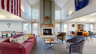 Located at 97 SW Rose Hill Cir in Topeka, Kansas, this single-floor community center offers a generously sized, open layout designed to accommodate a variety of social and gathering activities. The space is defined by soaring vaulted ceilings that create an airy, open ambiance throughout.

The welcoming lounge area serves as a central focal point, featuring a stone-surround fireplace with a warm wood mantel flanked by tall windows that flood the space with natural light. Comfortable seating arrangements invite relaxed conversation and leisure.

Adjacent to the lounge, a expansive dining and event hall provides ample room for large gatherings, furnished with multiple round tables and stackable chairs. The hall is illuminated by overhead fluorescent lighting and accented by vaulted ceilings, with windows along the perimeter offering outdoor views. A service counter area supports catering and event needs.

A bright, open activity room with large windows and plentiful natural light offers flexible small-group seating with modern cantilever chairs and small tables. Potted plants add a touch of warmth to this versatile space.

Additional areas include an entry foyer with a seating row and coat storage, private office or meeting rooms, and a library nook visible in the floor plan. Soft carpeting throughout contributes to a comfortable, community-oriented atmosphere well-suited for events, programs, and daily social activities.